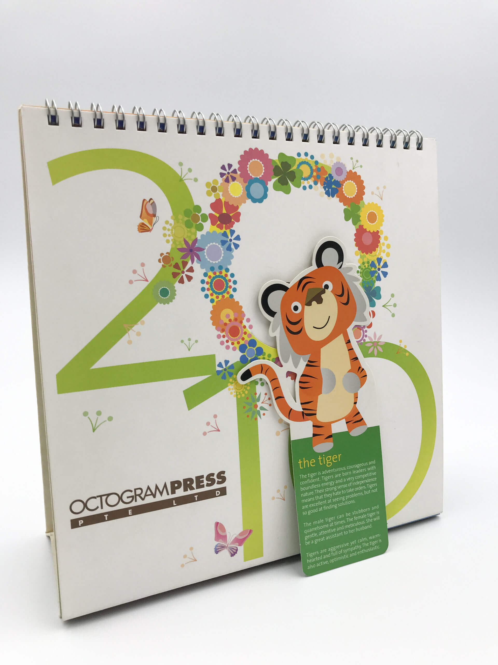 A custom-printed, spiral-binding desk calendar featuring a colorful floral design, a cute tiger illustration, and corporate branding for Octogram Press.