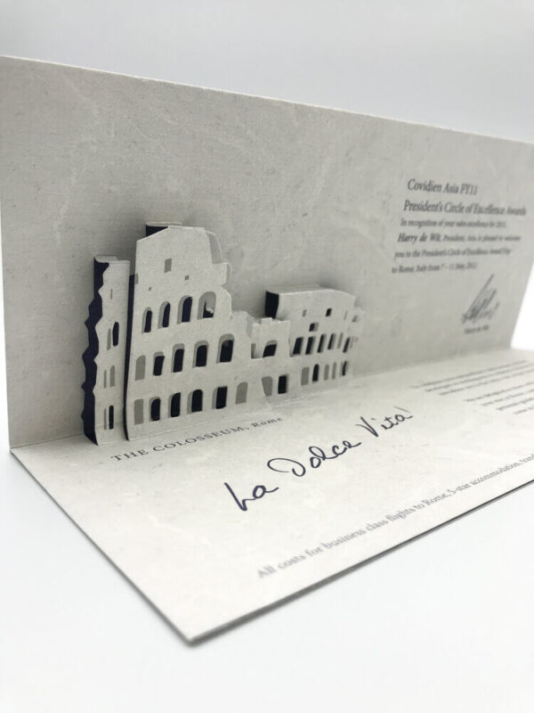 A custom corporate invitation featuring an intricate, multi-layered die-cut pop-up of the Roman Colosseum, crafted from textured grey paper for a President's Circle of Excellence award.