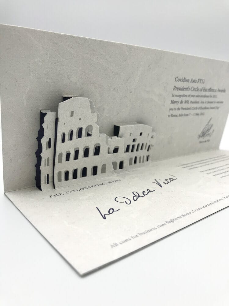 A custom corporate invitation featuring an intricate, multi-layered die-cut pop-up of the Roman Colosseum, crafted from textured grey paper for a President's Circle of Excellence award.