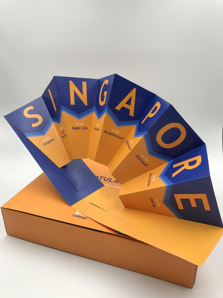 A creative promotional item featuring a die-cut, accordion-folded fan that forms the word 'SINGAPORE,' displayed in its coordinating orange gift box