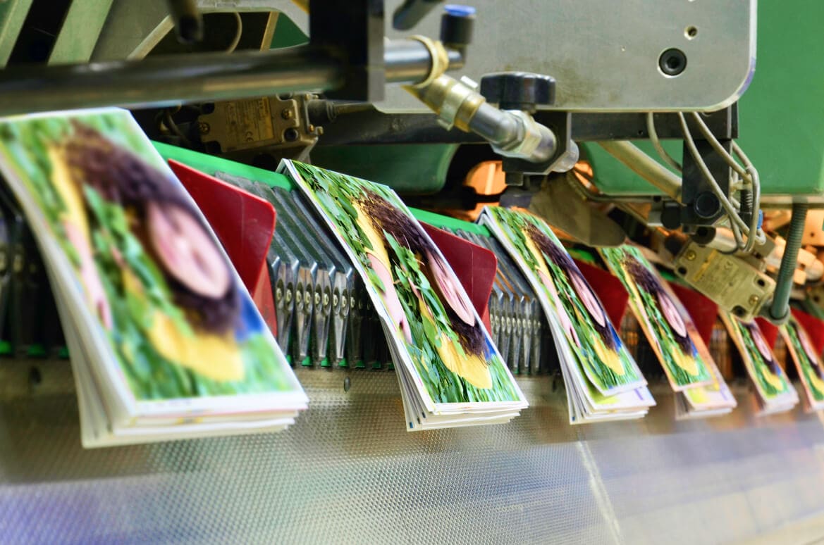 A close-up of a commercial offset printing press in Singapore, where newly printed, full-color brochures are being collated and stacked on a production line.