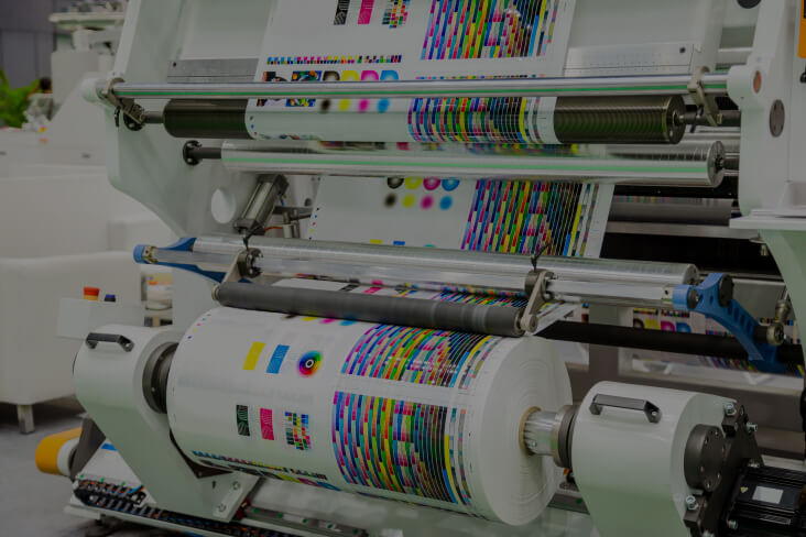 A large, industrial printing press in operation, feeding a wide roll of paper with vibrant CMYK color test patterns through its rollers.