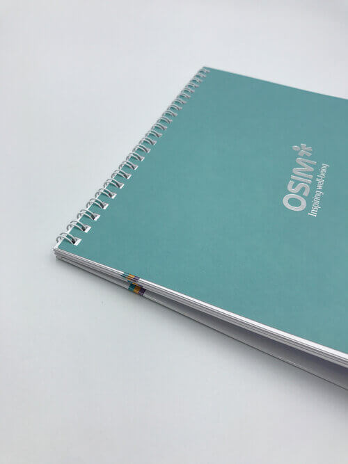 A close-up of a teal-covered notebook with durable silver spiral binding, featuring a custom silver foil logo for OSIM on a clean white background.