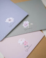 Three beautifully designed paper folders or notebooks are fanned out, showcasing their different pastel colors and minimalist floral designs on high-quality, textured paper