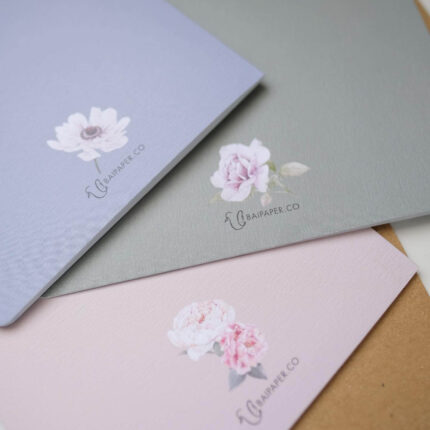 Three beautifully designed paper folders or notebooks are fanned out, showcasing their different pastel colors and minimalist floral designs on high-quality, textured paper