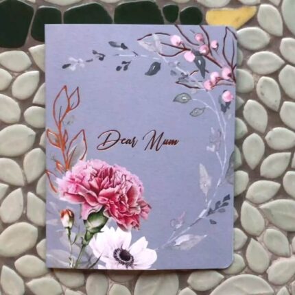 An artistic 'Dear Mum' greeting card with a lovely illustration of a carnation and foliage, accented with copper foil details, displayed on a textured, leaf-patterned tile background