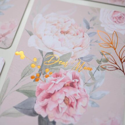 A close-up of a beautiful 'Dear Mum' greeting card featuring a soft pink watercolor floral design with elegant gold foil stamping on the text and leaf details.