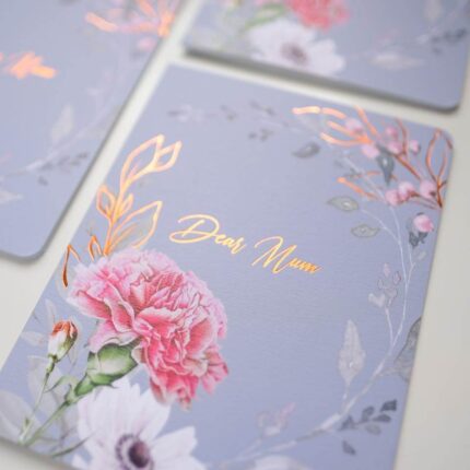 A set of elegant 'Dear Mum' greeting cards on a dusty blue background, featuring a pink carnation floral design accented with shiny rose gold foil on the lettering and botanical details.