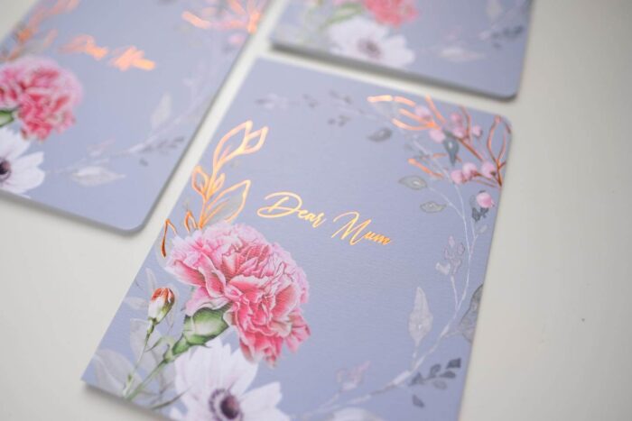 A set of elegant 'Dear Mum' greeting cards on a dusty blue background, featuring a pink carnation floral design accented with shiny rose gold foil on the lettering and botanical details.