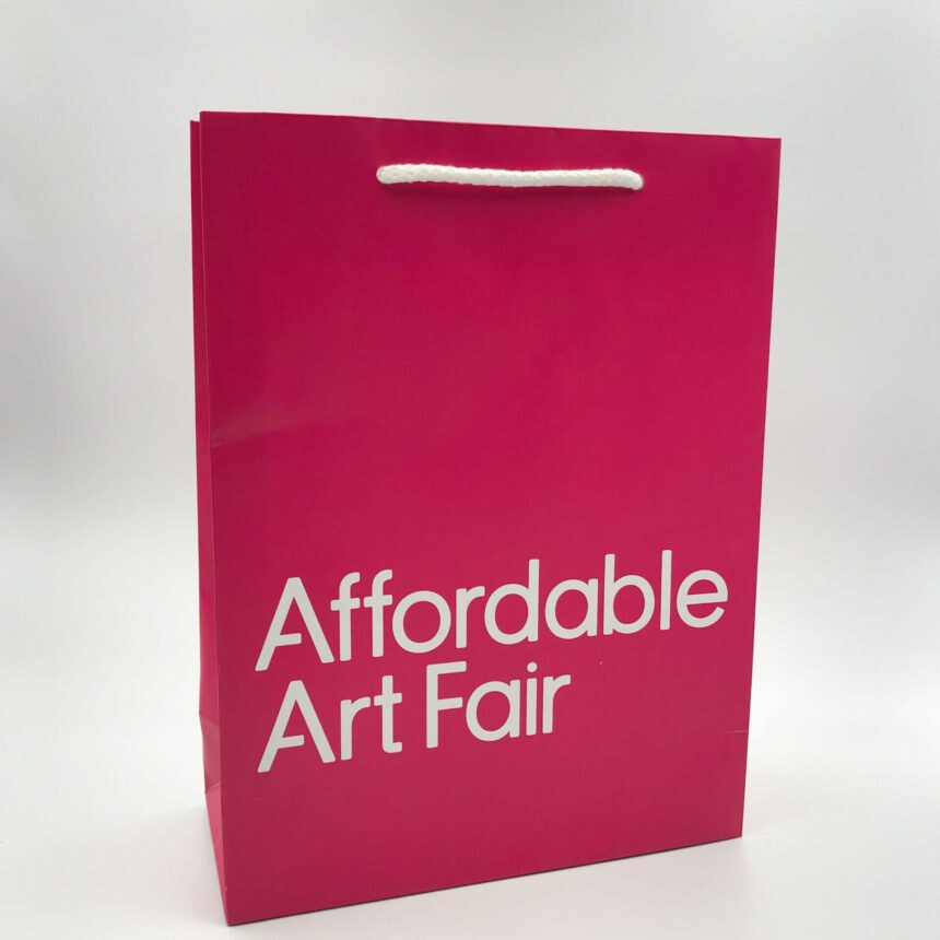 A bright pink, custom-printed paper bag for the 'Affordable Art Fair,' featuring the event title in bold white text and white rope handles.
