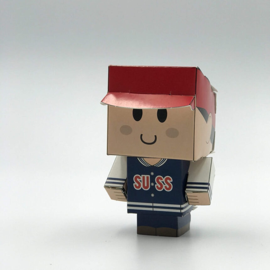 A custom-printed and die-cut papercraft toy of a character wearing a red cap and a blue 'SUSS' jacket, assembled and standing on a white background.