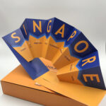 An intricately die-cut promotional fan that spells 'SINGAPORE,' with each letter highlighting a different aspect of the city, presented in a custom orange box.