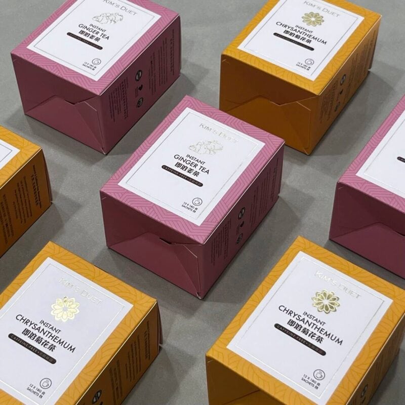 An arrangement of custom-printed boxes for Kim's Duet Instant Ginger Tea and Instant Chrysanthemum tea, showcasing the elegant pink and yellow product packaging with foil details.