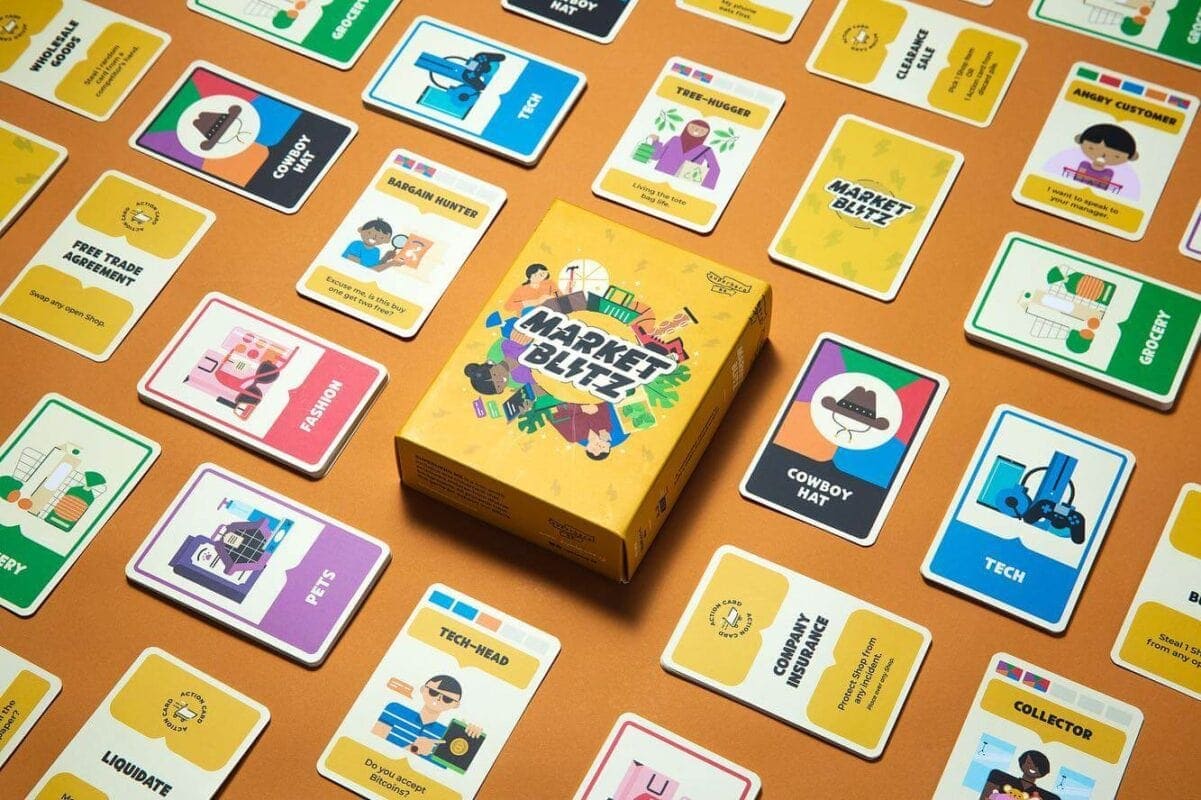 The 'Market Blitz' card game box is displayed on an orange surface, surrounded by a wide array of professionally printed, illustrated playing cards