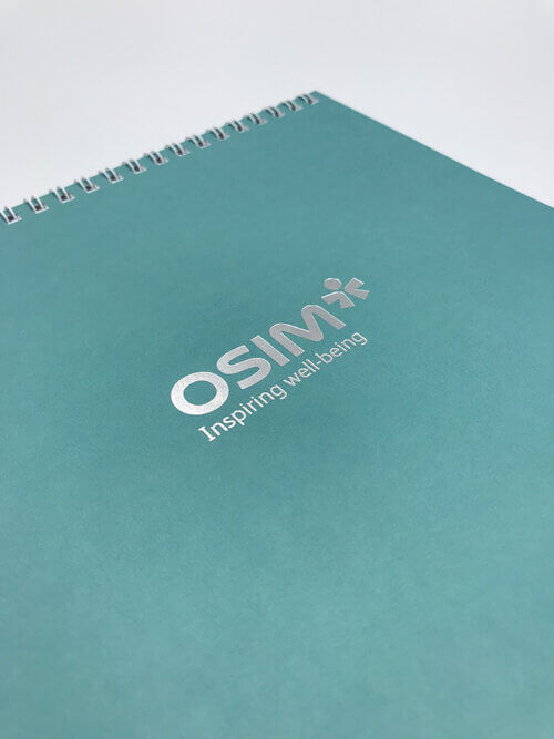 The cover of a teal, spiral-bound notebook for OSIM, featuring the company's logo and tagline 'Inspiring well-being' printed in elegant silver foil stamping.