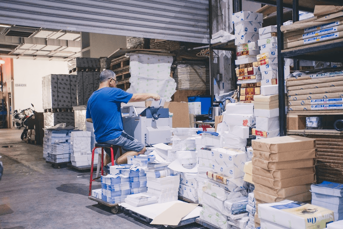 a man sitting on a stool operating a machine inside a busy printing warehouse surrounded by stacks of paper and packaged goods