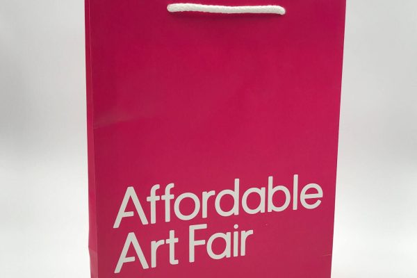 A bright pink, custom-printed paper bag for the 'Affordable Art Fair,' featuring the event title in bold white text and white rope handles.