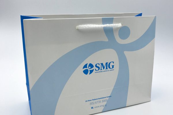 A custom-printed corporate paper bag for Singapore Medical Group (SMG), featuring their blue logo on a white background with rope handles and blue side gussets.