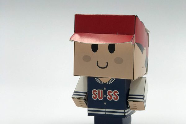 A custom-printed and die-cut papercraft toy of a character wearing a red cap and a blue 'SUSS' jacket, assembled and standing on a white background.