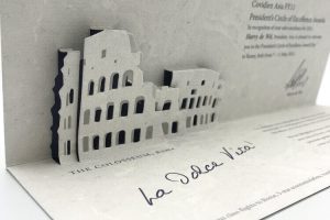 A custom corporate invitation featuring an intricate, multi-layered die-cut pop-up of the Roman Colosseum, crafted from textured grey paper for a President's Circle of Excellence award.