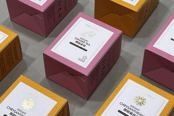 An arrangement of custom-printed boxes for Kim's Duet Instant Ginger Tea and Instant Chrysanthemum tea, showcasing the elegant pink and yellow product packaging with foil details.