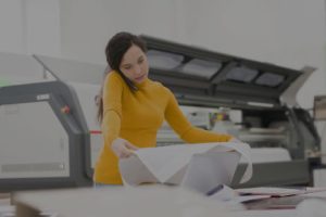 A print production specialist in a yellow shirt performs a quality assurance check, carefully inspecting a large printed proof while on the phone, with a large format printer visible in the background.
