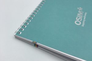 A close-up of a teal-covered notebook with durable silver spiral binding, featuring a custom silver foil logo for OSIM on a clean white background.