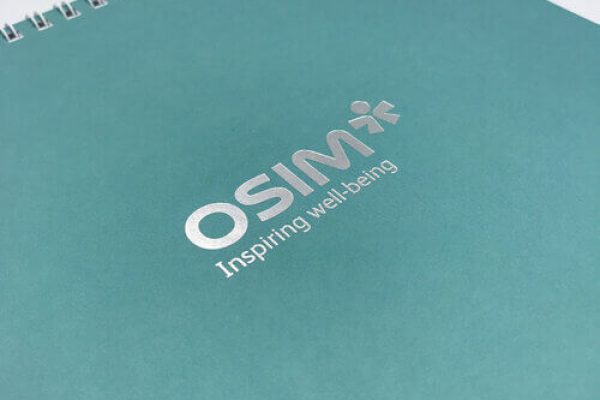 The cover of a teal, spiral-bound notebook for OSIM, featuring the company's logo and tagline 'Inspiring well-being' printed in elegant silver foil stamping.