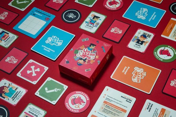 All the components of the 'Unleashed' card game are laid out on a red background, including the custom box, playing cards, attendance sheets, and die-cut tokens.